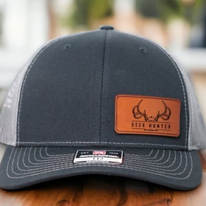 May include: A gray and white trucker hat with a brown leather patch that says "DEER HUNTER" with a stylized deer antler design.
