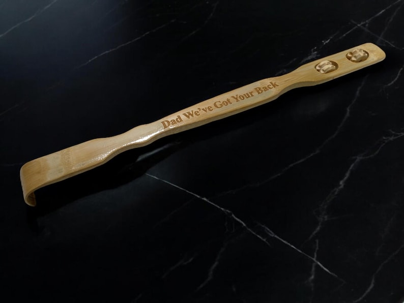 May include: A wooden back scratcher with a curved handle and two knobs. The scratcher is engraved with the text "Dad We've Got Your Back".