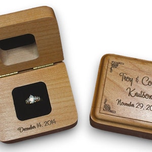 May include: A wooden ring box with a hinged lid and a velvet interior. The box is engraved with the names "Troy & Corinne Krutson" and the date "November 29, 2014". The lid of the box is open, revealing a ring inside.