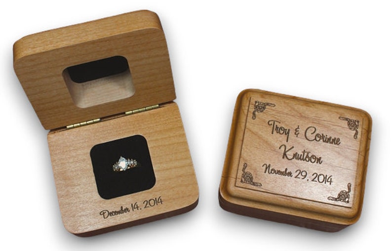 May include: A wooden ring box with a hinged lid and a velvet interior. The box is engraved with the names "Troy & Corinne Krutson" and the date "November 29, 2014". The lid of the box is open, revealing a ring inside.