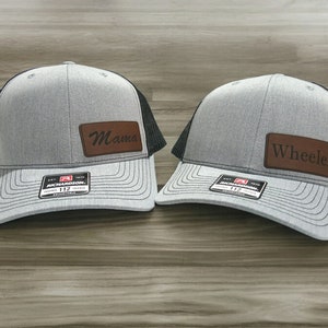 May include: Two gray trucker hats with black mesh backs and curved visors. One hat has a brown leather patch with the word "Mama", and the other has "Wheeler". Both hats have a Richardson 112 tag.