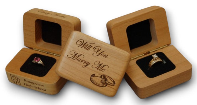 May include: Three wooden ring boxes. One box is engraved with "Will You Marry Me" and two rings. Another box is engraved with "Rangeview High School" and a red ring. The third box is empty.