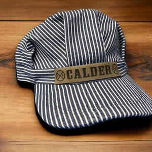 May include: A blue and white striped railroad engineer's cap with a brown leather patch that reads "RR CALDER" in black lettering.