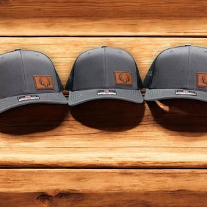 May include: Three gray and black trucker hats with a brown leather patch featuring a deer antler design.