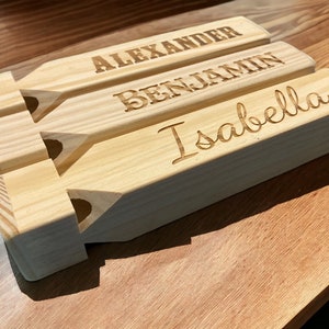 May include: Three personalized wooden name puzzles. Each puzzle is rectangular with a unique name engraved: Alexander, Benjamin, and Isabella. The wood has a natural, light color. The puzzles are displayed on a wooden surface.