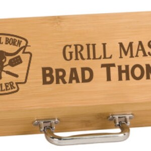 May include: A natural bamboo grilling tool set with a brown engraved design. The design features a bull skull with crossed grilling tools and the text "Natural Born Griller". The text "Grill Master Brad Thompson" is also engraved on the box.