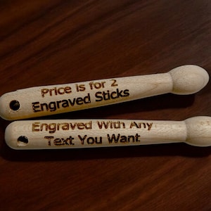 May include: Two wooden drumsticks with the text "Price is for 2 Engraved Sticks" and "Engraved With Any Text You Want" burned into the wood.
