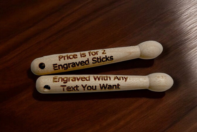 May include: Two wooden drumsticks with the text "Price is for 2 Engraved Sticks" and "Engraved With Any Text You Want" burned into the wood.