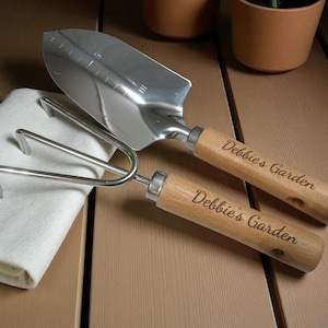 May include: A gardening set featuring a trowel and a hand fork with wooden handles. The trowel has measurement markings. The handles are engraved with "Debbie's Garden". A white cloth rests beside the tools.