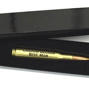 May include: A gold-colored bullet casing with the text "CARSON BECKER BEST MAN" engraved on it. The bullet casing is in a black velvet box.