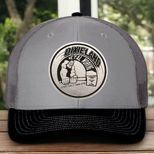 May include: A gray and black trucker hat with a silver metal patch that says "Dixieland Metal Working" and a cartoon image of a man working with a hammer. The patch also says "Open Daily 9AM-4PM".
