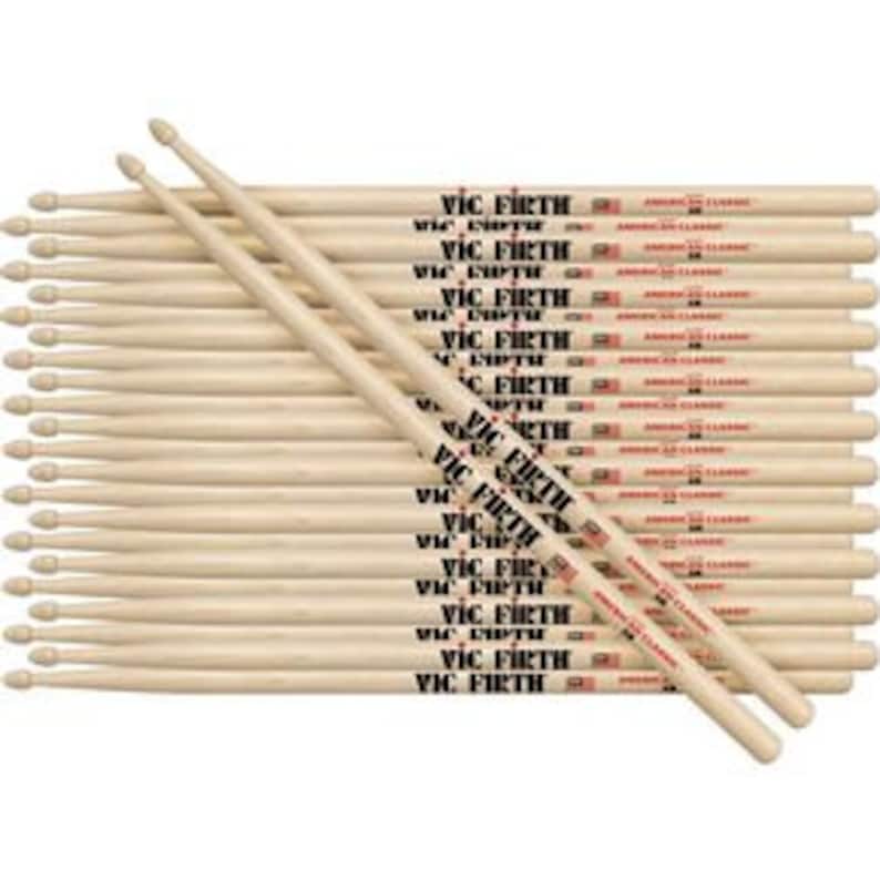 Puede incluir: Un conjunto de baquetas Vic Firth American Classic 5A. Las baquetas son de madera clara y presentan el logotipo de Vic Firth y un dise&ntilde;o de bandera estadounidense. Las baquetas est&aacute;n dispuestas en un mont&oacute;n ordenado.