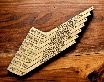 May include: Eight wooden hockey stick shaped signs with the text "Lady Swords BU 23-24" and player names and numbers. The signs are arranged in a fan shape on a wooden surface.