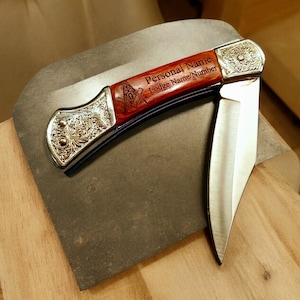 Puede incluir: Un cuchillo de bolsillo con mango de madera grabado con "Personal Name Lodge Name/Number" y un s&iacute;mbolo mas&oacute;nico. El cuchillo tiene una hoja de plata y un mango de color dorado.