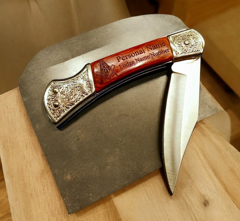 Puede incluir: Un cuchillo de bolsillo con mango de madera grabado con "Personal Name Lodge Name/Number" y un s&iacute;mbolo mas&oacute;nico. El cuchillo tiene una hoja de plata y un mango de color dorado.