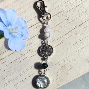 May include: A silver-toned metal keyring featuring a lobster clasp, two small white beads, a circular charm with an intricate openwork knot design, a single black textured bead, and a round pendant with a shimmering iridescent inlay.