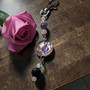 May include: A keyring featuring a silver-toned lobster clasp, a circular pendant with an iridescent shell inlay, and a series of beads including light pink, translucent green, purple, and a single textured black porous stone bead.