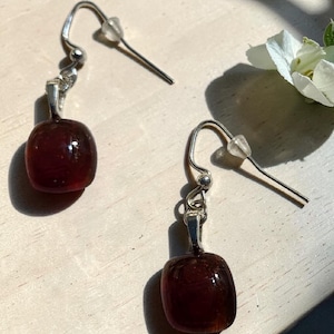 May include: A pair of dangle earrings with a silver hook and a rectangular, dark red, translucent bead. The earrings are displayed on a light-colored surface, with a white flower in the background.