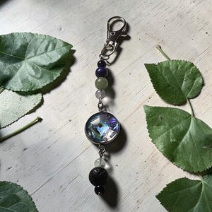 May include: A silver-toned metal keyring featuring a lobster clasp, a circular pendant with a multi-coloured iridescent inlay, and a series of beads including a black porous lava stone, a clear bead, a pale green bead, a dark blue bead, and a black bead.