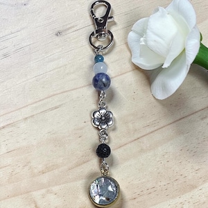 May include: A silver-toned metal keyring featuring a lobster clasp, a series of blue, white, and mottled purple beads, a silver-toned metal flower charm, a black porous bead, and a circular pendant with a shimmering iridescent inlay.