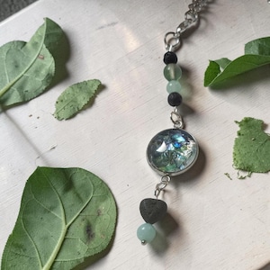 May include: A necklace featuring a silver-toned chain with a circular pendant containing iridescent blue and green fragments. The design includes black porous lava beads, translucent light green glass beads, and a small heart-shaped black stone.