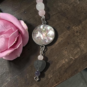 May include: A silver pendant necklace with a round, iridescent mother-of-pearl center. The necklace features pink and clear beads, a heart-shaped gray stone, and a purple bead. A pink rose is in the background.