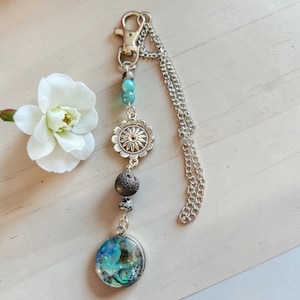 May include: A silver-toned keychain featuring a lobster clasp and a chain. The ornament includes a turquoise bead, a silver flower-shaped charm, a dark porous lava bead, a small speckled bead, and a circular pendant with an iridescent abalone inlay.