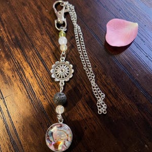 Peut inclure: Un porte-clés argenté composé d'un fermoir mousqueton, d'une fine chaîne et d'un arrangement vertical de perles. Le design comprend une petite perle vert citron, deux perles blanches translucides, une breloque fleur argentée, une perle de lave noire poreuse et un pendentif circulaire avec une incrustation de coquillage irisé multicolore.