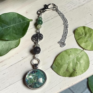 May include: A silver-toned keyring featuring a circular pendant with a textured teal and green inlay. The chain includes a silver tree-of-life charm, a porous black bead, and several smaller translucent and opaque green beads, finished with a lobster clasp.