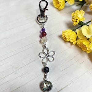 May include: A silver-toned metal keyring featuring a lobster clasp, a series of beads including purple, red, and translucent stones, a wire-formed flower charm, a black porous lava bead, and a circular pendant with an iridescent shell inlay.