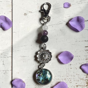 May include: A keychain featuring a silver-tone lobster clasp, a white bead, a purple bead, a black textured lava bead, a silver-tone flower-shaped charm, and a circular pendant with an iridescent, multi-colored abalone shell inlay.