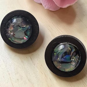 Peut inclure: Une paire de boucles d'oreilles à clous circulaires dotées d'un cadre en bois brun foncé entourant une incrustation centrale de nacre d'ormeau irisée aux tons tourbillonnants bleus, verts et violets. Les boucles ont une finition lisse et polie.