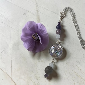 Può includere: Un ciondolo su catenina argentata con un elemento circolare riempito di frammenti di conchiglia iridescenti, arricchito da una serie di perline viola scuro, viola chiaro, bianco traslucido e una perla sferica grigio scuro testurizzata.