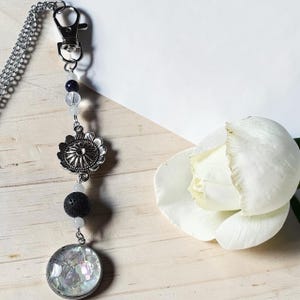 Peut inclure: Un porte-clés en métal argenté avec un fermoir mousqueton, une breloque fleur en argent, une perle de lave noire poreuse et un pendentif rond rempli de fragments de coquillages irisés, agrémenté de petites perles transparentes et bleu foncé.