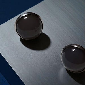Peut inclure: Deux objets sphériques gris foncé avec des bords argentés sont placés sur une surface grise, projetant des ombres. L'arrière-plan passe du gris au bleu foncé. Les objets semblent décoratifs ou fonctionnels, peut-être faisant partie d'un design plus large.