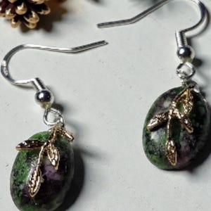Peut inclure: Boucles d'oreilles pendantes avec crochets argentés. Chaque boucle d'oreille présente une pierre ovale verte et violette avec un motif de feuille dorée. Les boucles d'oreilles sont présentées sur fond blanc avec des décorations de fête.