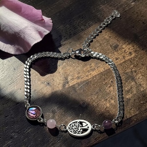 May include: A silver-toned chain bracelet featuring a circular charm with an iridescent purple inlay, a small pale pink bead, an oval charm depicting a tree, and a dark purple bead. The bracelet includes a lobster clasp and an adjustable extender chain.
