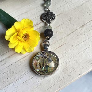 Può includere: Una collana con ciondolo color argento dotata di un pendente circolare riempito con frammenti di conchiglia iridescenti. La catena include una perla di lava nera porosa, un ciondolo color argento a forma di albero della vita e piccole perle verdi traslucide, vicino a un fiore giallo.