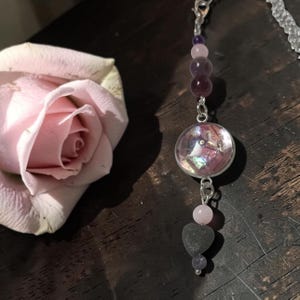 Può includere: Un ornamento decorativo da appendere con una catenina argentata, perline viola e rosa, un ciondolo circolare con un inserto iridescente scintillante, una piccola perla rosa chiaro e un ciondolo a forma di cuore grigio scuro in fondo.