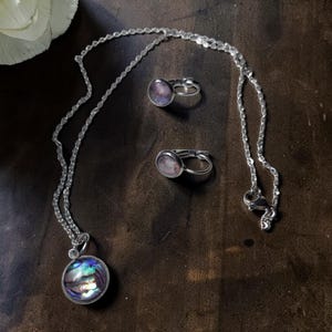 Könnte beinhalten: Eine silberne Halskette mit einem Anhänger mit einer runden, irisierenden Abalone-Schale. Zwei passende Silberringe mit einem lila-rosa Design. Das Schmuckset wird auf einer dunkelbraunen Oberfläche präsentiert.