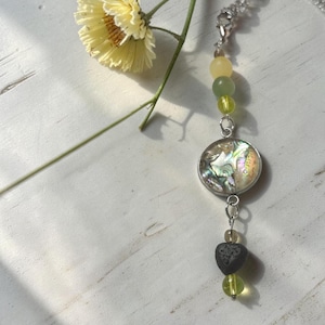 May include: A necklace featuring a silver-tone chain with a circular pendant containing iridescent abalone shell. The design includes a series of beads in shades of pale yellow, translucent green, and a dark gray heart-shaped bead at the base.