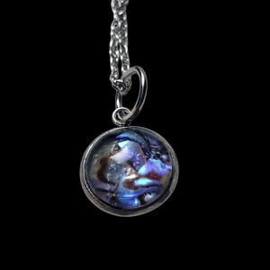 May include: A silver pendant necklace with a round, iridescent abalone shell pendant. The shell displays a mix of blue, purple, and brown colors. The chain is a delicate silver link. The background is solid black.
