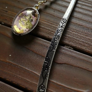 Peut inclure: Marque-page en métal argenté avec un charm ovale. Le charm contient des fleurs séchées et des paillettes dorées dans de la résine transparente. Le marque-page a un motif floral et est relié au charm par une fine chaîne.
