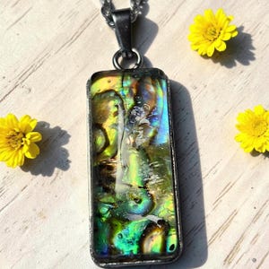 Può includere: Un ciondolo rettangolare con un inserto in conchiglia di abalone lucidata dai toni iridescenti verdi, blu e dorati, incastonato in una cornice di metallo scuro. Il ciondolo pende da una catena in metallo argentato, mostrando un motivo naturale vorticoso.
