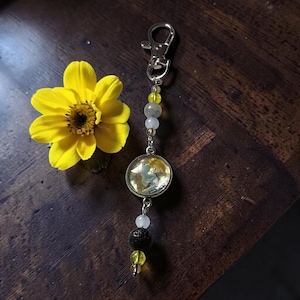 May include: A silver-toned metal keychain featuring a circular charm with a yellow and grey abstract design, accented by a series of small yellow, white, and grey beads, and one textured black porous bead, attached to a silver lobster clasp.