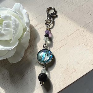May include: A keyring featuring a silver-tone lobster clasp, a round pendant with a blue and silver abstract metallic finish, and a series of beads including a black porous stone, a translucent pale green bead, a purple bead, and small clear spacers.