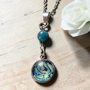 Peut inclure: Un collier argenté composé d'un pendentif rond avec une incrustation de coquille d'ormeau irisée, d'une perle ronde bleu sarcelle et d'une chaîne métallique. Le pendentif présente des tons tourbillonnants bleus, verts et nacrés.