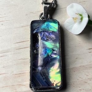 Può includere: Un ciondolo rettangolare caratterizzato da un inserto iridescente multicolore con sfumature di blu profondo, viola e verde brillante. Il ciondolo è racchiuso in una cornice di metallo argentato e include una contromaglia in metallo argentato.