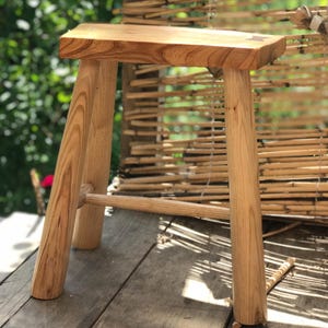 May include: A handcrafted wooden stool with a rectangular seat and four sturdy legs. The stool is made of light-colored wood, showcasing the natural grain. A horizontal support bar connects the legs. The stool is approximately 18 inches tall.