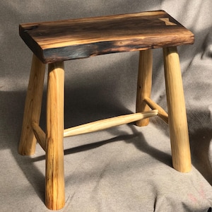 Puede incluir: Un taburete de madera hecho a mano con un asiento rectangular y cuatro patas resistentes. El asiento presenta un acabado marrón oscuro con detalles de madera más claros. Las patas son de madera clara natural, con una barra de soporte horizontal.